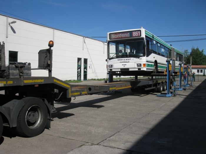 Der LKW-Tieflader fährt unter den angehobenen Gelenkobus.